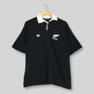 May include: A black short-sleeve rugby shirt featuring a white collar and a three-button placket. The chest displays a white embroidered Canterbury logo on the right and a white New Zealand All Blacks fern logo with text on the left side.