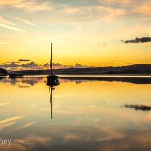 May include: A sailboat is moored in a still body of water at sunset. The sky is a vibrant orange and yellow, with clouds reflected in the water.