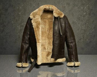 May include: A brown leather jacket with a tan shearling lining and collar. The jacket features a zip closure, side pockets, and a buckled belt at the waist. The cuffs are also lined with shearling.