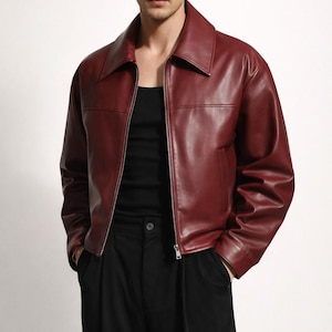 May include: A burgundy leather jacket with a collared neckline and a front zipper closure. The jacket is paired with a black tank top and black pants. The model is standing in front of a white background.