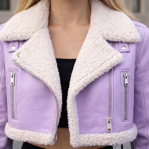 May include: A cropped lavender jacket with a white, fluffy shearling collar and trim. The jacket features a zipper closure and silver hardware. The style is reminiscent of a classic moto jacket, offering a chic and cozy look.