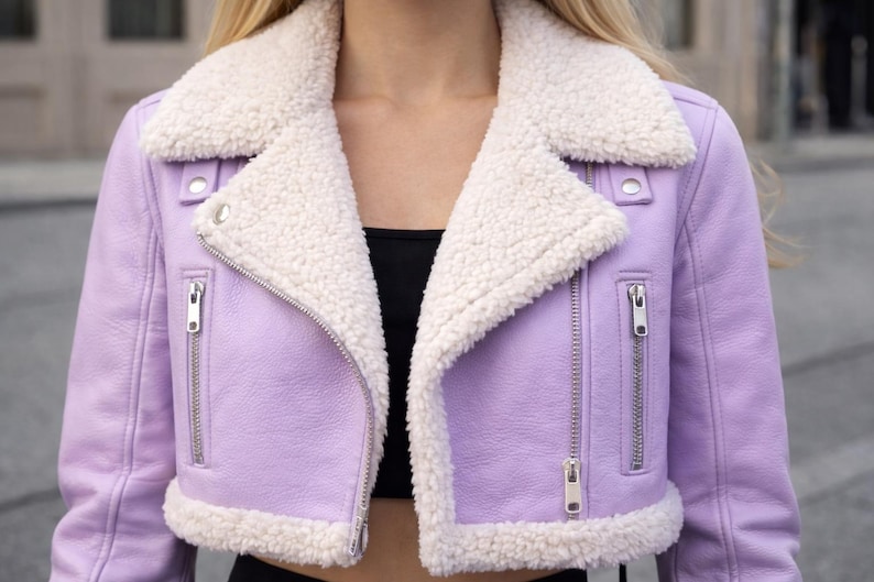 May include: A cropped lavender jacket with a white, fluffy shearling collar and trim. The jacket features a zipper closure and silver hardware. The style is reminiscent of a classic moto jacket, offering a chic and cozy look.