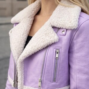 May include: A cropped lavender jacket with a white faux fur collar and trim. The jacket features a silver zipper closure and a classic biker jacket design. The soft, pastel color and plush trim create a stylish outerwear piece.