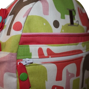 Op de afbeelding: Een roze, groene en bruine canvas rugzak met een ritssluiting en een roze en rode bies. De rugzak heeft een grote, geborduurde letter "F" aan de voorkant.