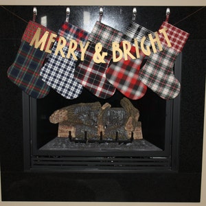 May include: Six plaid Christmas stockings hang from a fireplace mantel with a gold banner that reads "Merry & Bright".