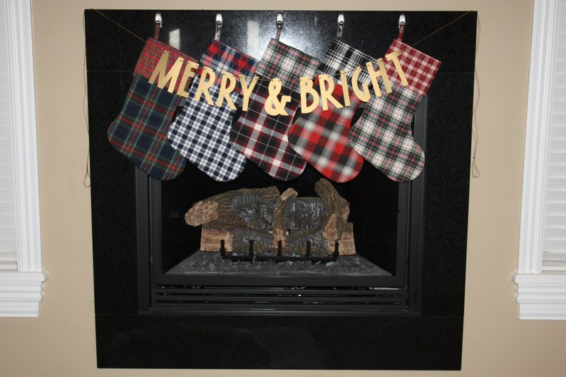 May include: Six plaid Christmas stockings hang from a fireplace mantel with a gold banner that reads "Merry & Bright".