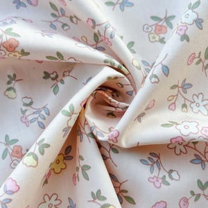 Könnte beinhalten: Nahaufnahme eines floralen Stoffes mit hellpfirsichfarbenem Hintergrund. Der Stoff weist ein sich wiederholendes Muster aus kleinen Blumen in Pastellfarben, darunter Rosa, Gelb und Blau, mit grünen Blättern auf. Der Stoff ist gefaltet und erzeugt Schatten und Tiefe.