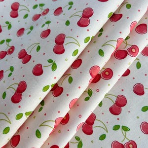 Könnte beinhalten: Nahaufnahme eines weißen Stoffes mit einem sich wiederholenden Muster aus roten Kirschen und grünen Blättern. Die Kirschen haben einen Aquarelleffekt, und der Stoff ist gefaltet, wodurch die Textur und das Design sichtbar werden.