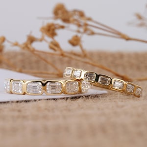 May include: Two gold rings with emerald-cut gemstones. The rings are set in a yellow gold band, with each ring featuring multiple clear, rectangular stones. The rings are displayed on a neutral background with dried floral accents.