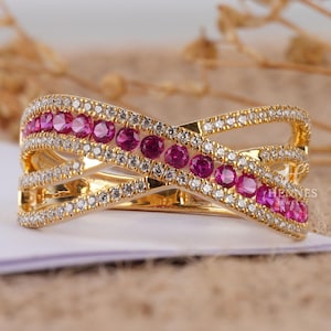May include: A gold-toned ring with an interwoven design. The ring features a row of pink gemstones and is encrusted with small, clear stones. The ring has a polished finish and is set against a neutral background. The ring is a piece of jewelry.