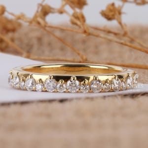May include: A gold band ring with a row of clear, round-cut gemstones. The ring is set on a white surface with a neutral background. The ring is a piece of jewelry.