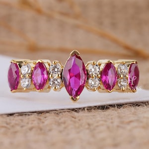 May include: A gold ring featuring marquise-cut ruby gemstones and small round white gemstones. The ruby gemstones are a vibrant pink color, and the ring has a delicate, elegant design. The ring is set on a white surface.