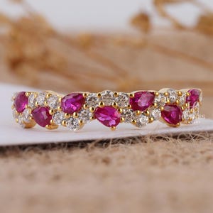 May include: A gold ring featuring alternating pear-shaped ruby gemstones and round white diamonds. The gemstones are set in a double row, creating a sparkling and elegant design. The ring is set on a neutral background.