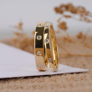 May include: Two gold rings with multiple small, clear gemstones set into the band. The rings are positioned together, showcasing their polished, reflective surface. The rings are on a white surface with a neutral background.