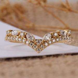May include: A gold-colored ring with a V-shaped design. The ring is adorned with numerous small, round, and marquise-cut clear gemstones. The ring is set on a white surface with a blurred beige background. The text "HENNES JEWELRY" is visible on the right side of the ring.