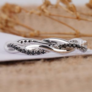 May include: A silver-colored ring with a twisted design, featuring a row of small black stones. The ring has a polished finish and is set against a neutral background. The ring is a piece of jewelry.