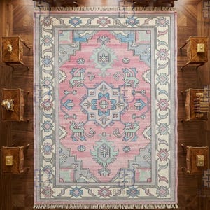 K&ouml;nnte beinhalten: Draufsicht auf einen rechteckigen, rosafarbenen Teppich mit einem detaillierten geometrischen Muster in Blau-, Grau- und Wei&szlig;t&ouml;nen. Der Teppich hat einen Fransenrand und ist von Holzm&ouml;beln und kleinen St&uuml;hlen umgeben.