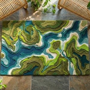 May include: A rectangular rug with a topographical design in shades of green, blue, and white. The rug's pattern resembles a landscape with flowing lines and varying textures. The rug is surrounded by plants and wicker chairs.