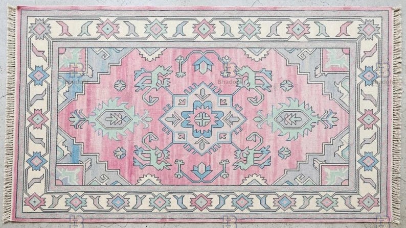 K&ouml;nnte beinhalten: Rechteckiger Teppich mit rosa Mittelfeld und cremefarbenem Rand. Geometrische Muster in Blau, Grau und Mintgr&uuml;n. Die Kanten haben Fransen. Der Teppich ist f&uuml;r die Inneneinrichtung im traditionellen Stil konzipiert.