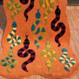 Puede incluir: Una alfombra naranja texturizada con un patrón repetitivo de serpientes en varios colores, incluyendo amarillo, verde, azul, negro y morado. Las serpientes están representadas en un movimiento sinuoso sobre la superficie de la alfombra.