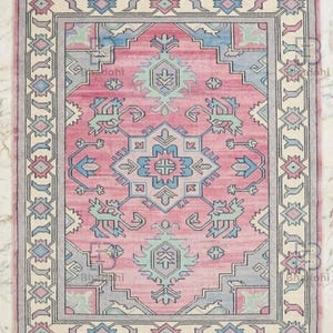 K&ouml;nnte beinhalten: Rechteckiger rosa Teppich mit einem zentralen geometrischen Design in Blau und Gr&uuml;n. Der Teppich hat einen cremefarbenen Rand mit geometrischen Mustern und Fransen an den Kanten. Geeignet f&uuml;r ein Wohn- oder Schlafzimmer.