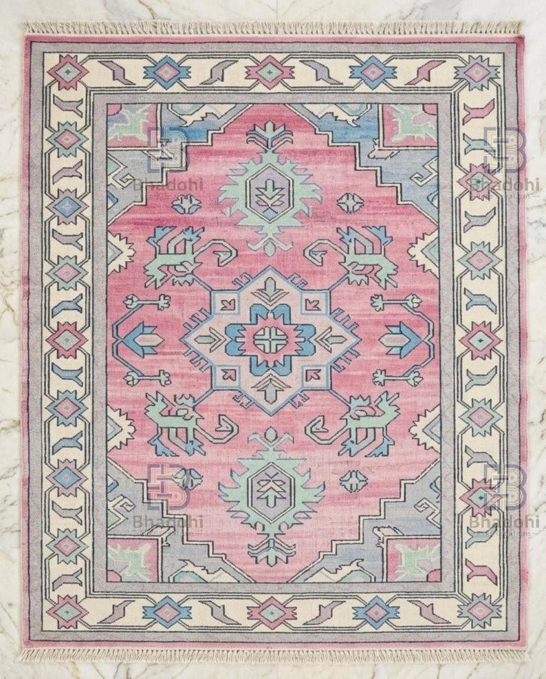 K&ouml;nnte beinhalten: Rechteckiger rosa Teppich mit einem zentralen geometrischen Design in Blau und Gr&uuml;n. Der Teppich hat einen cremefarbenen Rand mit geometrischen Mustern und Fransen an den Kanten. Geeignet f&uuml;r ein Wohn- oder Schlafzimmer.