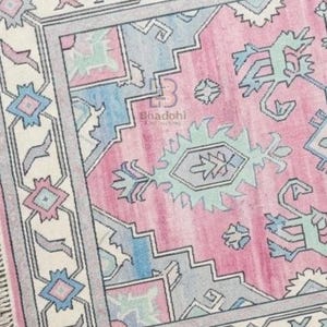 K&ouml;nnte beinhalten: Rechteckiger Teppich in Rosa, Blau und Creme. Der Teppich hat ein zentrales rosa Feld mit geometrischen und floralen Mustern in Blau, Creme und Grau. Der Rand ist cremefarben mit sich wiederholenden geometrischen Formen.