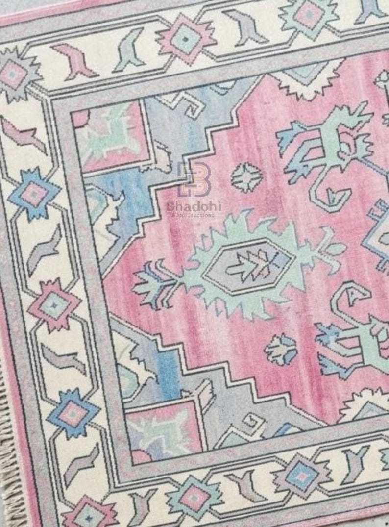 K&ouml;nnte beinhalten: Rechteckiger Teppich in Rosa, Blau und Creme. Der Teppich hat ein zentrales rosa Feld mit geometrischen und floralen Mustern in Blau, Creme und Grau. Der Rand ist cremefarben mit sich wiederholenden geometrischen Formen.
