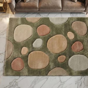 May include: A rectangular area rug with a pebble design. The rug features large, rounded shapes in shades of beige, peach, and gray, set against a green background. The rug is placed on a marble floor.