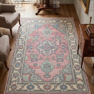 K&ouml;nnte beinhalten: Ein rechteckiger Teppich mit einem rosa Mittelfeld und einem detaillierten Rand. Der Teppich weist geometrische Muster in Blau-, Grau- und Cremet&ouml;nen auf. Der Teppich hat einen Fransenbesatz am unteren Rand und liegt auf einem Holzboden.