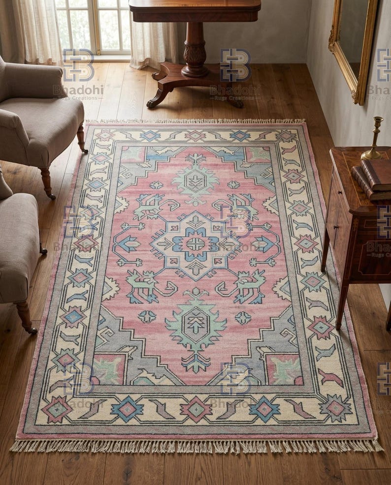 K&ouml;nnte beinhalten: Ein rechteckiger Teppich mit einem rosa Mittelfeld und einem detaillierten Rand. Der Teppich weist geometrische Muster in Blau-, Grau- und Cremet&ouml;nen auf. Der Teppich hat einen Fransenbesatz am unteren Rand und liegt auf einem Holzboden.