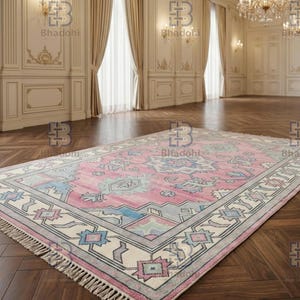 K&ouml;nnte beinhalten: Ein rechteckiger, rosa-grauer Teppich mit einem detaillierten geometrischen Muster. Der Teppich hat einen cremefarbenen Rand mit blauen, rosa- und grauen Akzenten. Der Teppich hat Fransen an den kurzen Enden und liegt auf einem dunklen Holzboden.