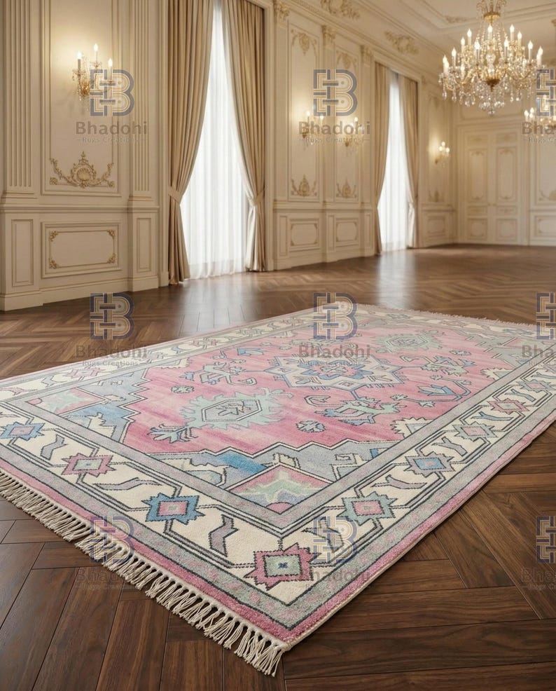 K&ouml;nnte beinhalten: Ein rechteckiger, rosa-grauer Teppich mit einem detaillierten geometrischen Muster. Der Teppich hat einen cremefarbenen Rand mit blauen, rosa- und grauen Akzenten. Der Teppich hat Fransen an den kurzen Enden und liegt auf einem dunklen Holzboden.
