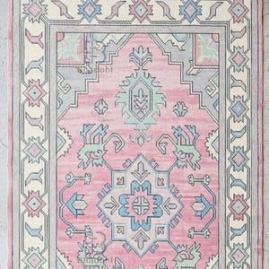 K&ouml;nnte beinhalten: Ein rechteckiger rosa Teppich mit einem komplizierten geometrischen Muster. Der Teppich hat ein zentrales Design in Rosa, Blau und Gr&uuml;n, das von einem cremefarbenen und grauen Rand umgeben ist. Die Kanten haben einen Fransen.