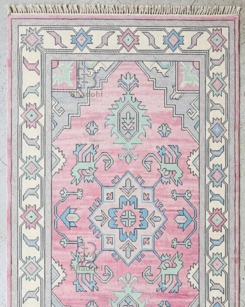 K&ouml;nnte beinhalten: Ein rechteckiger rosa Teppich mit einem komplizierten geometrischen Muster. Der Teppich hat ein zentrales Design in Rosa, Blau und Gr&uuml;n, das von einem cremefarbenen und grauen Rand umgeben ist. Die Kanten haben einen Fransen.