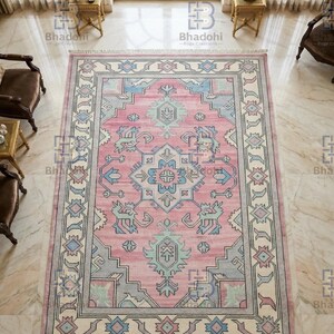K&ouml;nnte beinhalten: Ein rechteckiger, rosafarbener Teppich mit einem zentralen geometrischen Muster in Blau-, Gr&uuml;n- und Graut&ouml;nen. Der Teppich hat einen cremefarbenen Rand mit sich wiederholenden geometrischen Mustern und Fransen. Er liegt auf einem Marmorboden.