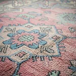 K&ouml;nnte beinhalten: Nahaufnahme eines handgewebten Teppichs mit detailliertem geometrischem Muster. Der Teppich hat eine Farbpalette aus Rosa, Blau und Creme. Die Textur des Teppichs ist sichtbar und zeigt die komplizierte Webart und das Design.