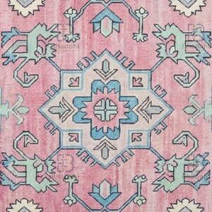K&ouml;nnte beinhalten: Nahaufnahme eines rosa Teppichs mit einem zentralen geometrischen Design in Hellblau, T&uuml;rkis und Off-White. Der Teppich weist ein kompliziertes Muster mit sich wiederholenden floralen und stilisierten Tiermotiven auf und erzeugt eine Vintage-&Auml;sthetik.