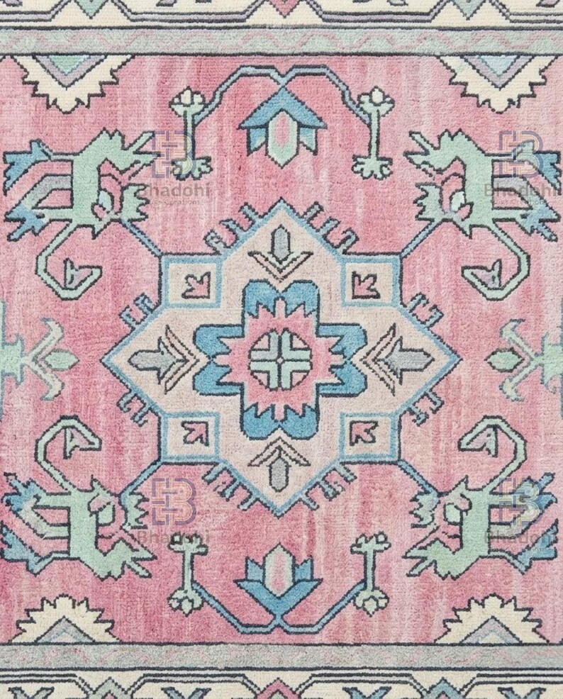 K&ouml;nnte beinhalten: Nahaufnahme eines rosa Teppichs mit einem zentralen geometrischen Design in Hellblau, T&uuml;rkis und Off-White. Der Teppich weist ein kompliziertes Muster mit sich wiederholenden floralen und stilisierten Tiermotiven auf und erzeugt eine Vintage-&Auml;sthetik.