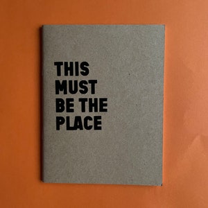 Può includere: Un quaderno marrone con il testo "THIS MUST BE THE PLACE" stampato in nero.
