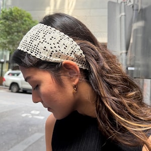 Könnte beinhalten: Ein cremefarbenes, gehäkeltes Stirnband mit geometrischem Muster. Das Stirnband wird auf dem Kopf getragen. Die Person trägt goldene Ohrringe und ein schwarzes Oberteil. Der Hintergrund ist eine Stadtstraße.