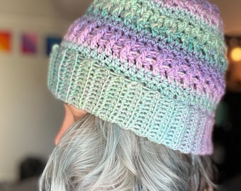May include: A handmade crochet beanie in pastel colors. The hat features a ribbed brim in shades of light green and blue, transitioning to a body of lavender, green, and blue. The crochet stitch creates a textured appearance, perfect for cold weather.