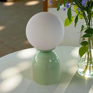 May include: A modern table lamp with a spherical white shade atop a light green base. The lamp has a rounded, bulbous shape and sits on a white surface. A vase of flowers is in the background.