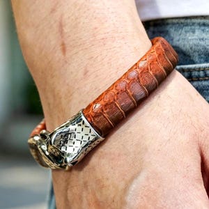 May include: A brown leather bracelet with a reptile skin texture. The bracelet features a silver-coloured metal clasp with a decorative design. The bracelet is worn on a wrist, with blue jeans visible in the background.
