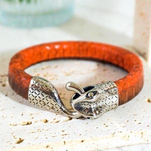 Puede incluir: Pulsera de cuero naranja y marrón con cierre de cabeza de serpiente plateada. La pulsera presenta una banda de cuero texturizado y un diseño detallado de cabeza de serpiente. Un accesorio elegante.