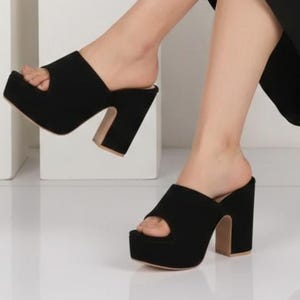 May include: Black platform sandals with a peep-toe design. The sandals have a thick heel and a suede-like texture. The image also shows a person wearing a black dress and holding a silver clutch. Two pairs of beige shoes are displayed in the background.