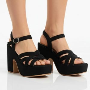 May include: Black platform sandals with crisscross straps across the foot and an ankle strap with a silver buckle. The chunky heel and platform sole are black, with a tan-colored sole.