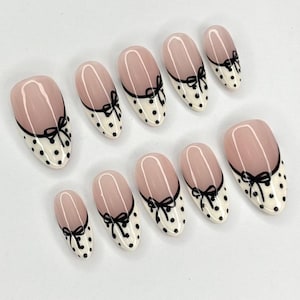 May include: Set of ten oval-shaped false nails. Each nail features a light pink base with a black bow design and a white polka dot pattern. The nails are arranged on a white surface.