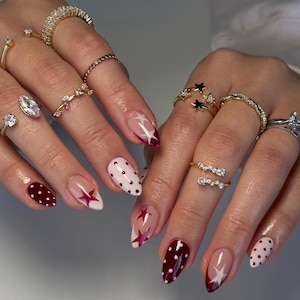 May include: Close-up of hands with various gold rings and manicured nails. The nails feature designs with burgundy tips, white polka dots, and star patterns. The rings include diamond and gemstone accents.