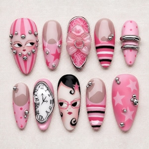 May include: A collection of pink and nude-colored artificial nails with various designs. Some nails feature stripes, stars, and faces, while others have embellishments like silver studs and a bow. One nail displays a clock face.